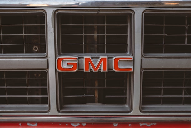 A close-up of a vehicle grille featuring the bold red letters 'GMC' prominently in the center. The grille is metallic with a grid pattern and shows some signs of wear.