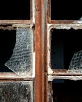 A close-up of a broken window with rusty metal frames. The glass is shattered, missing pieces, and covered in dust. The texture of the glass shows a honeycomb pattern, and the frame shows signs of wear and decay.
