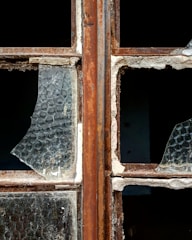 A close-up of a broken window with rusty metal frames. The glass is shattered, missing pieces, and covered in dust. The texture of the glass shows a honeycomb pattern, and the frame shows signs of wear and decay.