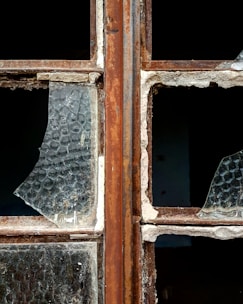 A close-up of a broken window with rusty metal frames. The glass is shattered, missing pieces, and covered in dust. The texture of the glass shows a honeycomb pattern, and the frame shows signs of wear and decay.