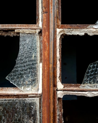 A close-up of a broken window with rusty metal frames. The glass is shattered, missing pieces, and covered in dust. The texture of the glass shows a honeycomb pattern, and the frame shows signs of wear and decay.