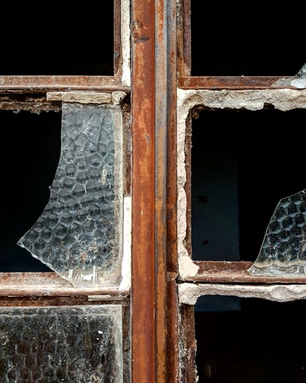 A close-up of a broken window with rusty metal frames. The glass is shattered, missing pieces, and covered in dust. The texture of the glass shows a honeycomb pattern, and the frame shows signs of wear and decay.