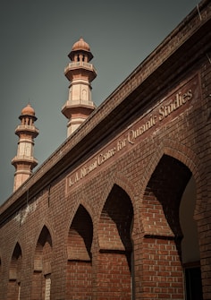 A brick building with ornate architectural details featuring pointed arches and two taller minarets. The structure has a sign labeled 'K.A. Nizami Centre for Quranic Studies' indicating its academic or religious significance.