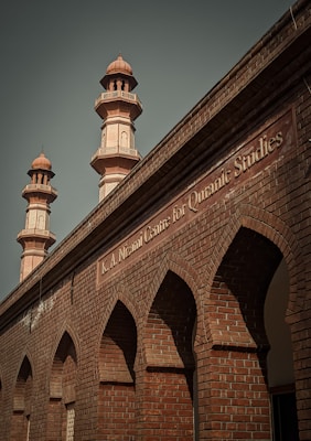 A brick building with ornate architectural details featuring pointed arches and two taller minarets. The structure has a sign labeled 'K.A. Nizami Centre for Quranic Studies' indicating its academic or religious significance.