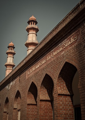 A brick building with ornate architectural details featuring pointed arches and two taller minarets. The structure has a sign labeled 'K.A. Nizami Centre for Quranic Studies' indicating its academic or religious significance.