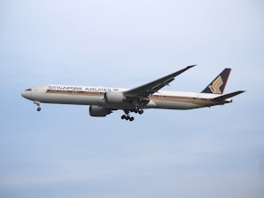 A large commercial airplane from Singapore Airlines is flying in the sky, showcasing its distinctive livery and logo on the fin. The aircraft appears to be in mid-flight with landing gear retracted, set against a clear blue sky.