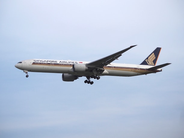 A large commercial airplane from Singapore Airlines is flying in the sky, showcasing its distinctive livery and logo on the fin. The aircraft appears to be in mid-flight with landing gear retracted, set against a clear blue sky.