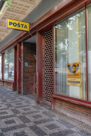 A sidewalk outside a building with large windows featuring vertical blinds and a sign on the window displaying the logo of 'Česk&aacute; Po&scaron;ta'. The building facade is constructed with brown and red brick, and a prominent yellow and blue 'PO&Scaron;TA' sign hangs above the entrance.