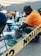 A person wearing a cap and orange shirt is working with several open cans of paint, mixing green paint with a brush. Various paint cans with colors like yellow and blue are placed around, and plastic covers the ground. Yellow caution tape is visible in the foreground.