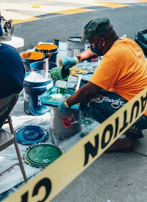 A person wearing a cap and orange shirt is working with several open cans of paint, mixing green paint with a brush. Various paint cans with colors like yellow and blue are placed around, and plastic covers the ground. Yellow caution tape is visible in the foreground.