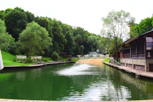 A serene lakeside scene surrounded by lush green trees. The water is calm and has a deep green hue, reflecting the dense forest. To the right, there's a wooden dock attached to a rustic building with large windows. Lounge chairs and canopied sunbeds line the shore, suggesting a relaxing atmosphere. At the far end, there is a sandy beach area with more vegetation in the background.