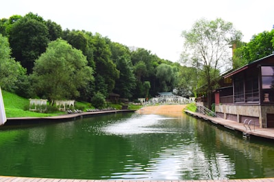 A serene lakeside scene surrounded by lush green trees. The water is calm and has a deep green hue, reflecting the dense forest. To the right, there's a wooden dock attached to a rustic building with large windows. Lounge chairs and canopied sunbeds line the shore, suggesting a relaxing atmosphere. At the far end, there is a sandy beach area with more vegetation in the background.