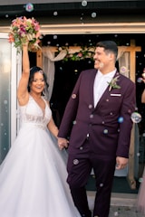 A newlywed couple is depicted joyfully exiting a building. The bride, dressed in a white gown, holds a bouquet of flowers aloft, while the groom, wearing a maroon suit, smiles at her. Bubbles are floating around them, adding a whimsical touch to the scene. In the background, there is floral decor above the doorway, suggesting a wedding celebration.