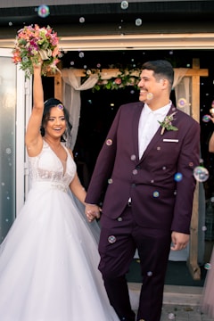 A newlywed couple is depicted joyfully exiting a building. The bride, dressed in a white gown, holds a bouquet of flowers aloft, while the groom, wearing a maroon suit, smiles at her. Bubbles are floating around them, adding a whimsical touch to the scene. In the background, there is floral decor above the doorway, suggesting a wedding celebration.