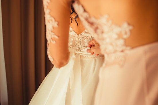 A bride is being helped with the back of her lace wedding dress, featuring intricate floral patterns and a satin ribbon being tied. The warm lighting creates an intimate and delicate atmosphere.