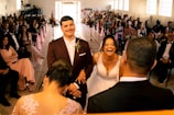 A wedding ceremony with the bride and groom standing together at the altar. The bride is laughing joyously while the groom, wearing a maroon suit, smiles gently. The scene includes guests seated on both sides, dressed in formal attire, with pink decorations and flowers lining the aisle.