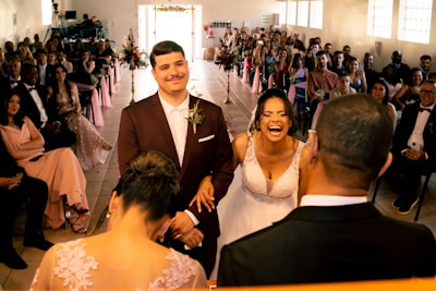 A wedding ceremony with the bride and groom standing together at the altar. The bride is laughing joyously while the groom, wearing a maroon suit, smiles gently. The scene includes guests seated on both sides, dressed in formal attire, with pink decorations and flowers lining the aisle.