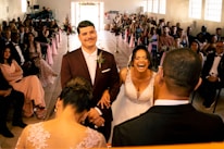 A wedding ceremony with the bride and groom standing together at the altar. The bride is laughing joyously while the groom, wearing a maroon suit, smiles gently. The scene includes guests seated on both sides, dressed in formal attire, with pink decorations and flowers lining the aisle.