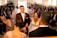 A wedding ceremony with the bride and groom standing together at the altar. The bride is laughing joyously while the groom, wearing a maroon suit, smiles gently. The scene includes guests seated on both sides, dressed in formal attire, with pink decorations and flowers lining the aisle.