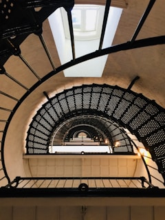 A spiraling metal staircase is viewed from below, creating an intricate pattern of curves and lines. The stairs curve upward leading to a bright window at the top, allowing natural light to pour in. The design features small perforations in the treads, adding texture and complexity to the geometric composition.