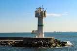 A tall white lighthouse with a green stripe stands on a rocky pier extending into a calm sea. Several birds are perched on top of the lighthouse. In the background, ships can be seen on the horizon under a clear blue sky.