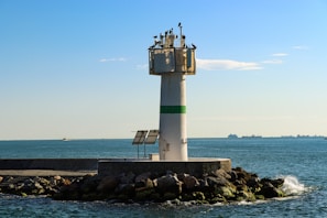 A tall white lighthouse with a green stripe stands on a rocky pier extending into a calm sea. Several birds are perched on top of the lighthouse. In the background, ships can be seen on the horizon under a clear blue sky.