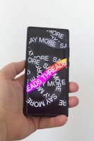A hand holding a smartphone with a screen displaying overlapping text in white saying 'SAY MORE' multiple times. There is a vibrant gradient text 'THREADS' in the foreground.