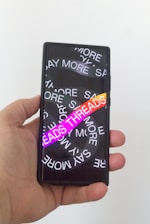 A hand holding a smartphone with a screen displaying overlapping text in white saying 'SAY MORE' multiple times. There is a vibrant gradient text 'THREADS' in the foreground.