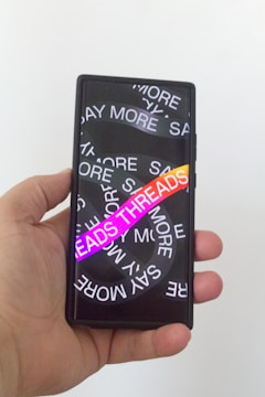 A hand holding a smartphone with a screen displaying overlapping text in white saying 'SAY MORE' multiple times. There is a vibrant gradient text 'THREADS' in the foreground.