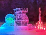 An illuminated ice sculpture featuring an astronaut with a smiling face on the left. The sculpture glows with a combination of blue, pink, and red lights. A rocket-shaped ice sculpture stands on the right-hand side, also illuminated by colored lights. Trees and snow can be seen in the dark background.