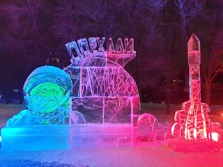 An illuminated ice sculpture featuring an astronaut with a smiling face on the left. The sculpture glows with a combination of blue, pink, and red lights. A rocket-shaped ice sculpture stands on the right-hand side, also illuminated by colored lights. Trees and snow can be seen in the dark background.