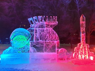 An illuminated ice sculpture featuring an astronaut with a smiling face on the left. The sculpture glows with a combination of blue, pink, and red lights. A rocket-shaped ice sculpture stands on the right-hand side, also illuminated by colored lights. Trees and snow can be seen in the dark background.