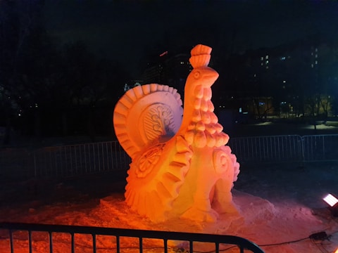 A large, intricately carved snow sculpture resembling a turkey is illuminated by warm, orange lights. The sculpture exhibits detailed feathers and a textured body, placed in an outdoor setting surrounded by metal barriers with buildings visible in the background.