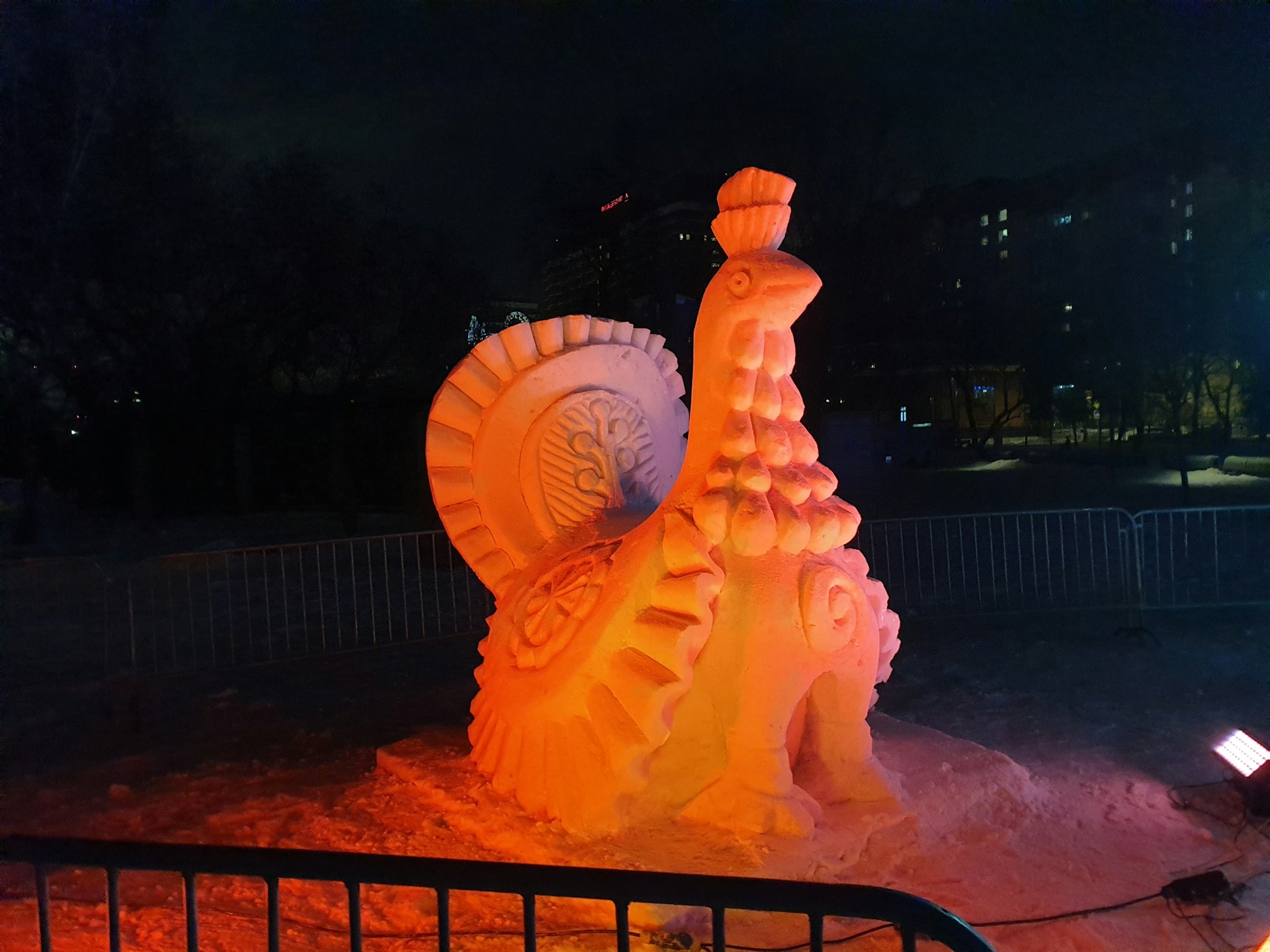 A large, intricately carved snow sculpture resembling a turkey is illuminated by warm, orange lights. The sculpture exhibits detailed feathers and a textured body, placed in an outdoor setting surrounded by metal barriers with buildings visible in the background.