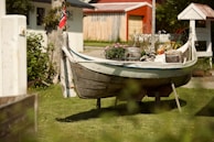 An old wooden boat is displayed on a grassy lawn, repurposed as a decorative planter filled with flowers and plants. A small national flag is attached to the boat, and nearby, a quaint white cottage with a red roof provides a backdrop. The scene is bathed in warm sunlight, and the area is surrounded by lush greenery and vibrant flowers.