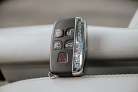 A Range Rover key fob rests on a light-colored leather surface. The fob features various buttons including lock, unlock, headlights, and hazard warning.