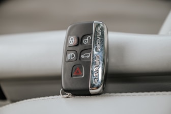 A Range Rover key fob rests on a light-colored leather surface. The fob features various buttons including lock, unlock, headlights, and hazard warning.