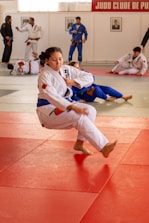 A martial arts dojo features several practitioners in white and blue gi uniforms engaged in training on red and gray mats. The central figure is performing a martial arts technique, surrounded by others practicing and discussing techniques.