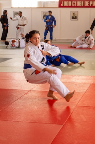 A martial arts dojo features several practitioners in white and blue gi uniforms engaged in training on red and gray mats. The central figure is performing a martial arts technique, surrounded by others practicing and discussing techniques.