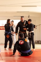 Five people wearing martial arts uniforms are gathered together in a dojo, smiling and interacting with each other. The environment is well-lit, with visible mats on the floor. The mood appears lighthearted and friendly.