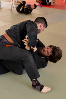 Two men are practicing Brazilian Jiu-Jitsu on a mat in a gym setting, wearing traditional gis with belts. The focus seems to be on grappling techniques with one person trying to lock the other in a submission hold. Other individuals are seen in the background engaged in similar activities.