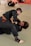 Two men are practicing Brazilian Jiu-Jitsu on a mat in a gym setting, wearing traditional gis with belts. The focus seems to be on grappling techniques with one person trying to lock the other in a submission hold. Other individuals are seen in the background engaged in similar activities.