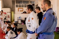 A group of individuals dressed in martial arts gis, gathered in a dojo. Most are seated, attentively listening, while one person in a blue gi stands prominently. The setting appears focused and disciplined, with some written notes visible on a bulletin board in the background.