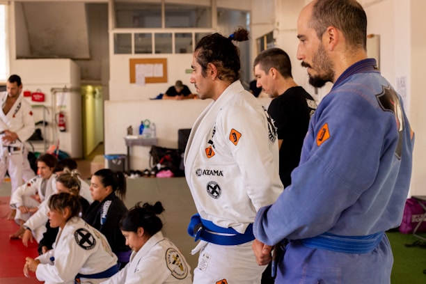 A group of individuals dressed in martial arts gis, gathered in a dojo. Most are seated, attentively listening, while one person in a blue gi stands prominently. The setting appears focused and disciplined, with some written notes visible on a bulletin board in the background.