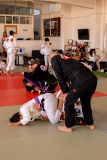 Participants in a Brazilian Jiu-Jitsu class practicing grappling techniques on a mat. They are wearing traditional gi uniforms and include a mixture of belts, indicating different skill levels. The setting is a spacious dojo with onlookers and other students in the background.