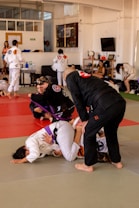 Participants in a Brazilian Jiu-Jitsu class practicing grappling techniques on a mat. They are wearing traditional gi uniforms and include a mixture of belts, indicating different skill levels. The setting is a spacious dojo with onlookers and other students in the background.