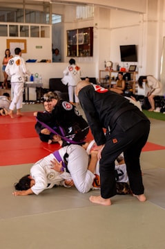 Participants in a Brazilian Jiu-Jitsu class practicing grappling techniques on a mat. They are wearing traditional gi uniforms and include a mixture of belts, indicating different skill levels. The setting is a spacious dojo with onlookers and other students in the background.