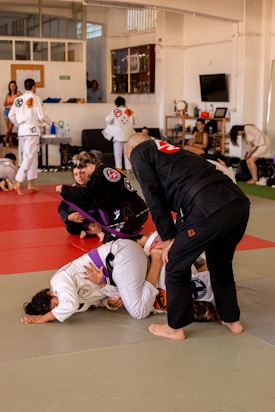 Participants in a Brazilian Jiu-Jitsu class practicing grappling techniques on a mat. They are wearing traditional gi uniforms and include a mixture of belts, indicating different skill levels. The setting is a spacious dojo with onlookers and other students in the background.