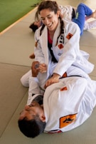 Two people are practicing Brazilian Jiu-Jitsu on a mat, wearing white gis with various patches. One person, smiling with braided hair, is in a top position, appearing to demonstrate a technique on the other person lying on their back.