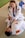 Two people are practicing Brazilian Jiu-Jitsu on a mat, wearing white gis with various patches. One person, smiling with braided hair, is in a top position, appearing to demonstrate a technique on the other person lying on their back.